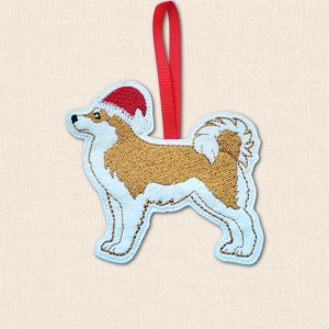 Golden and pale embroidered Akita wearing a red Santa hat, finished with a red hanging ribbon, on a cream fabric background.