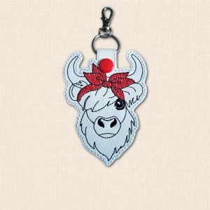 White faux leather Highland cow keyring with black embroidered shaggy hair, curved horns, a red bandana bow and metal lobster clasp.