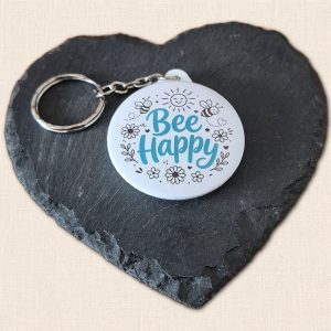 Round white 'Bee Happy' keyring with turquoise lettering, black bee and flower illustrations, a smiling sun and a silver-coloured split ring, displayed on a slate heart.