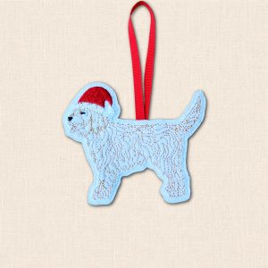 Standing Cockapoo embroidered felt decoration with a raised tail, red Santa hat and red hanging ribbon, displayed on cream fabric.