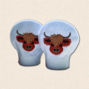 Pair of white felt LED tealight covers with embroidered Highland cow faces, brown shaggy fringes and curved horns.