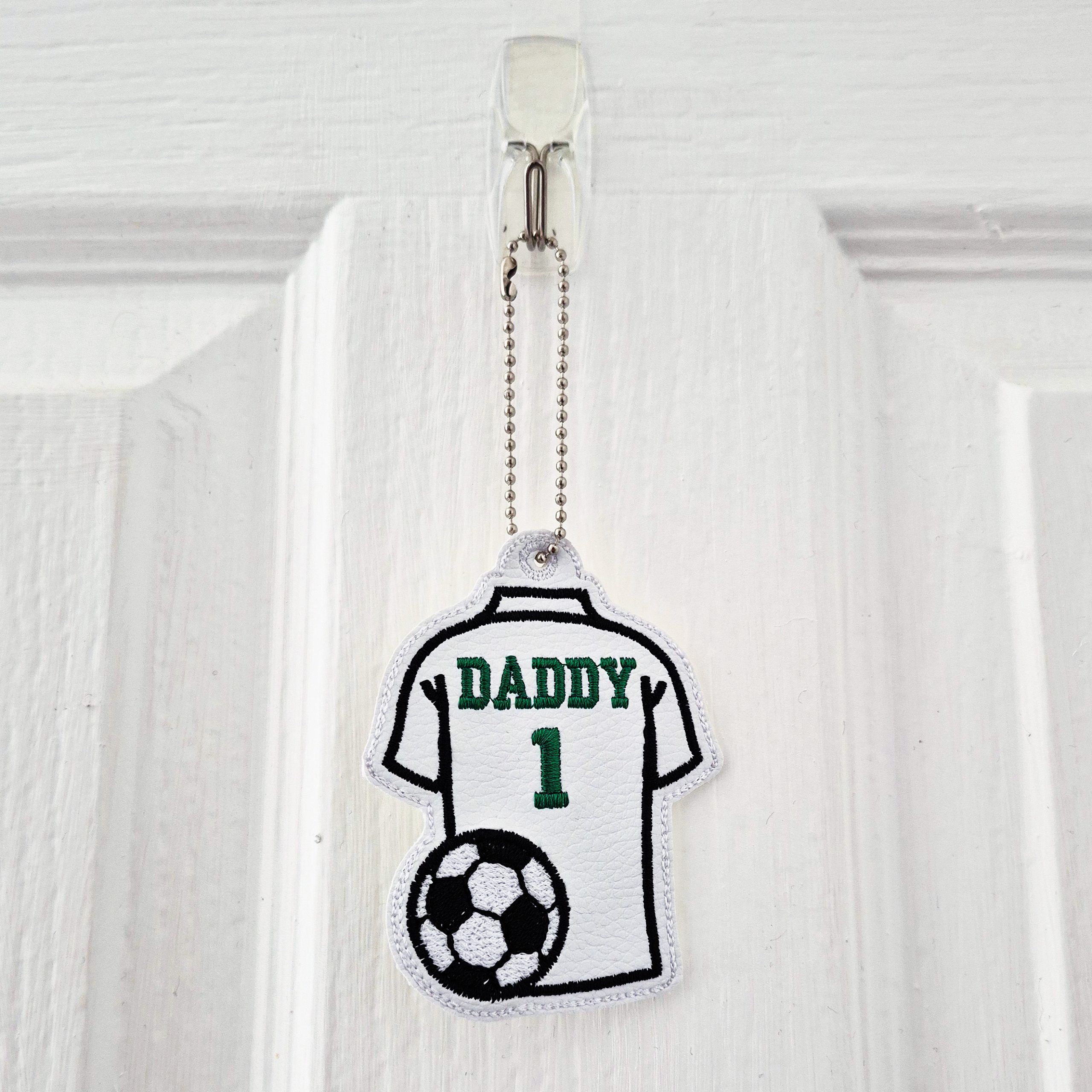 'Daddy 1' football shirt charm hanging from a hook on a white door by its metal ball chain.
