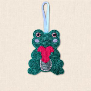 Green embroidered felt frog holding a bright pink heart, with a smiling face and ribbon hanging loop on a cream background.