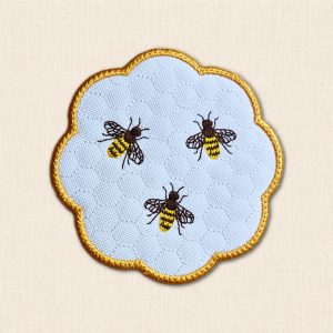 White candle coaster featuring three embroidered honey bees, subtle honeycomb stitching and a golden yellow scalloped border.