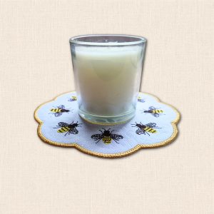Glass candle jar displayed on a white embroidered honey bee candle mat with golden yellow scalloped edging.