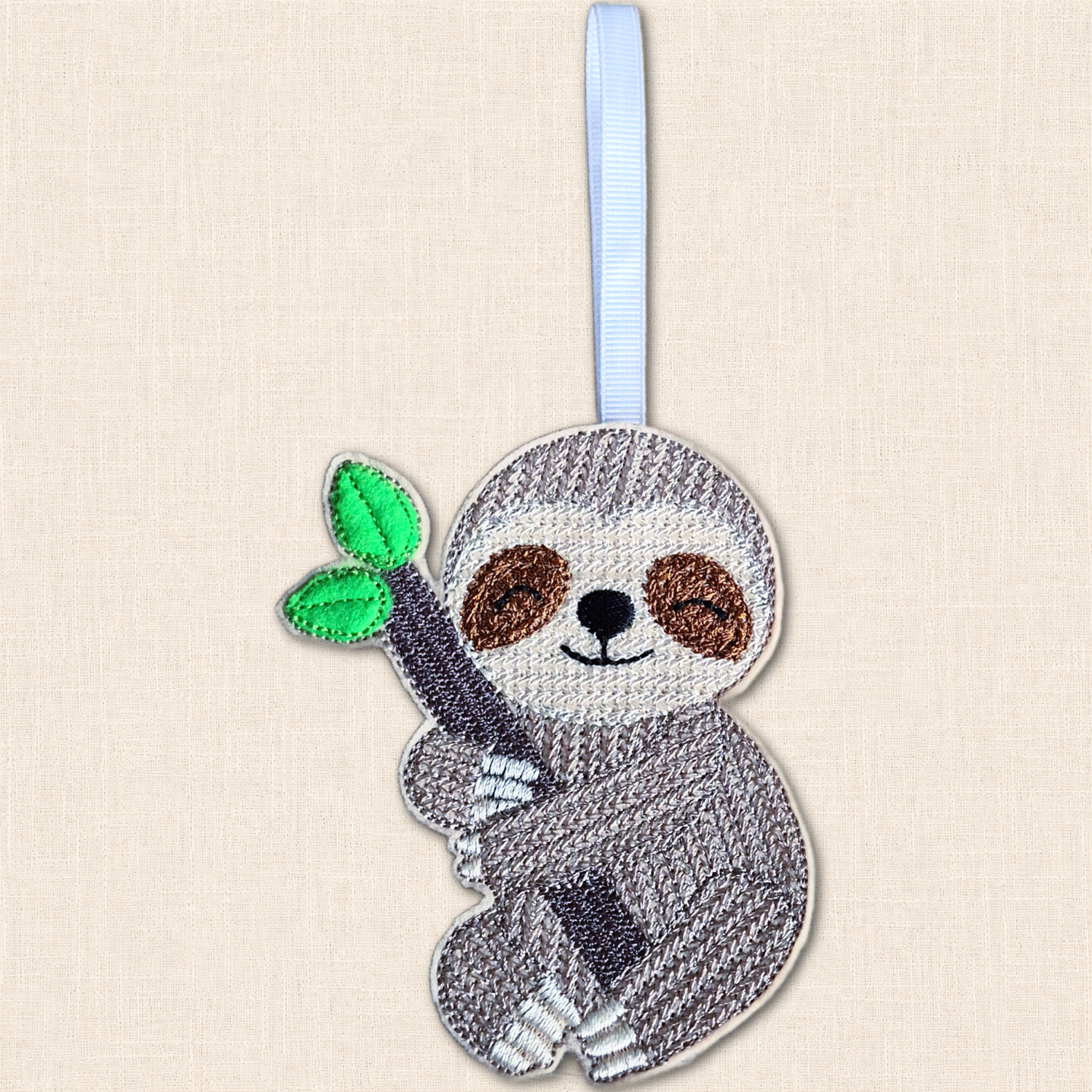 Close-up of an embroidered felt sloth holding a branch with two green leaves, showing knit-stitch detail, brown eye patches and a smiling face.