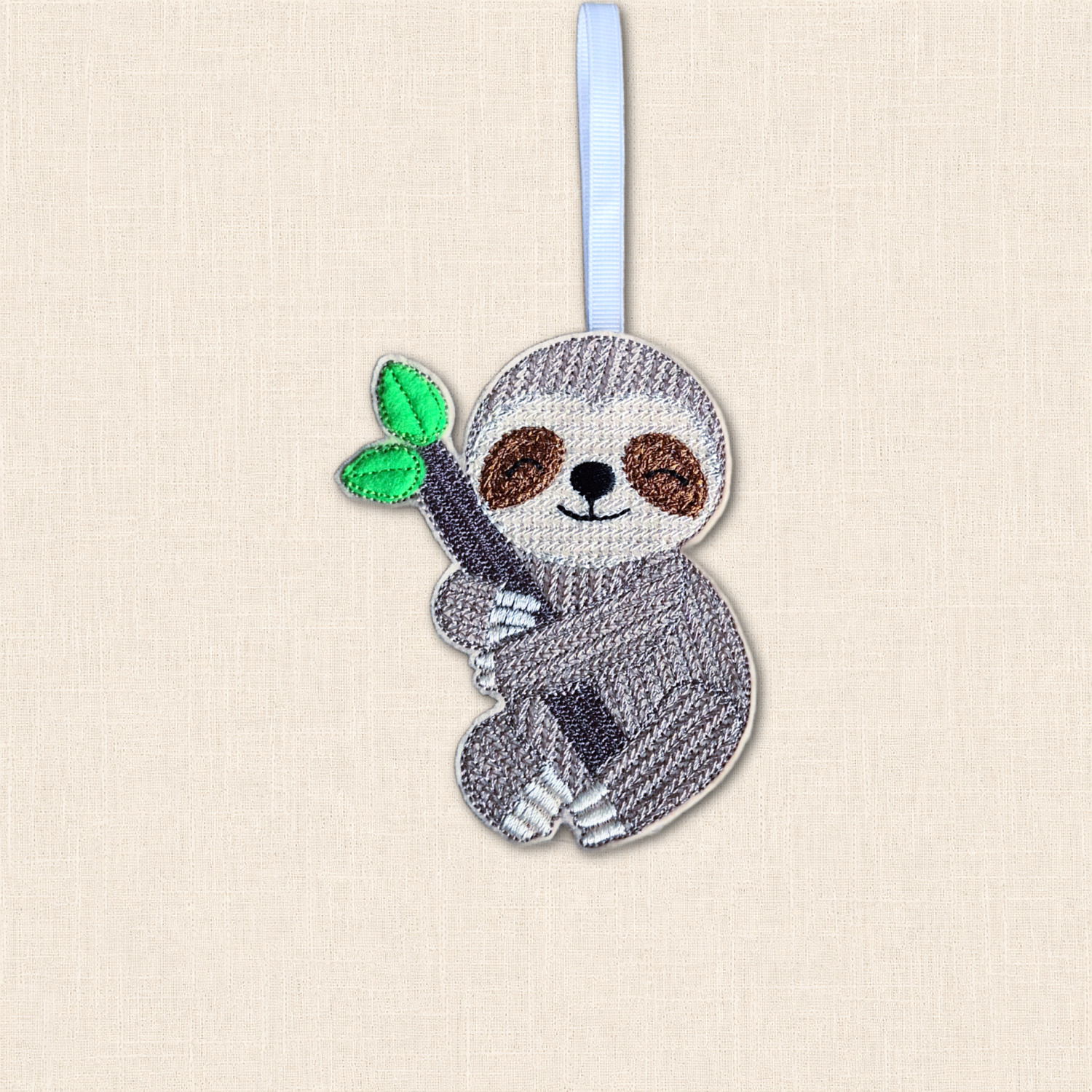 Smiling embroidered felt sloth holding a leafy branch, with a ribbon hanging loop against a cream background.