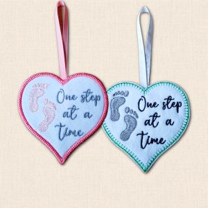 Two 'One Step at a Time' felt heart hangers with embroidered footprints, showing pink satin stitch and green blanket stitch borders.