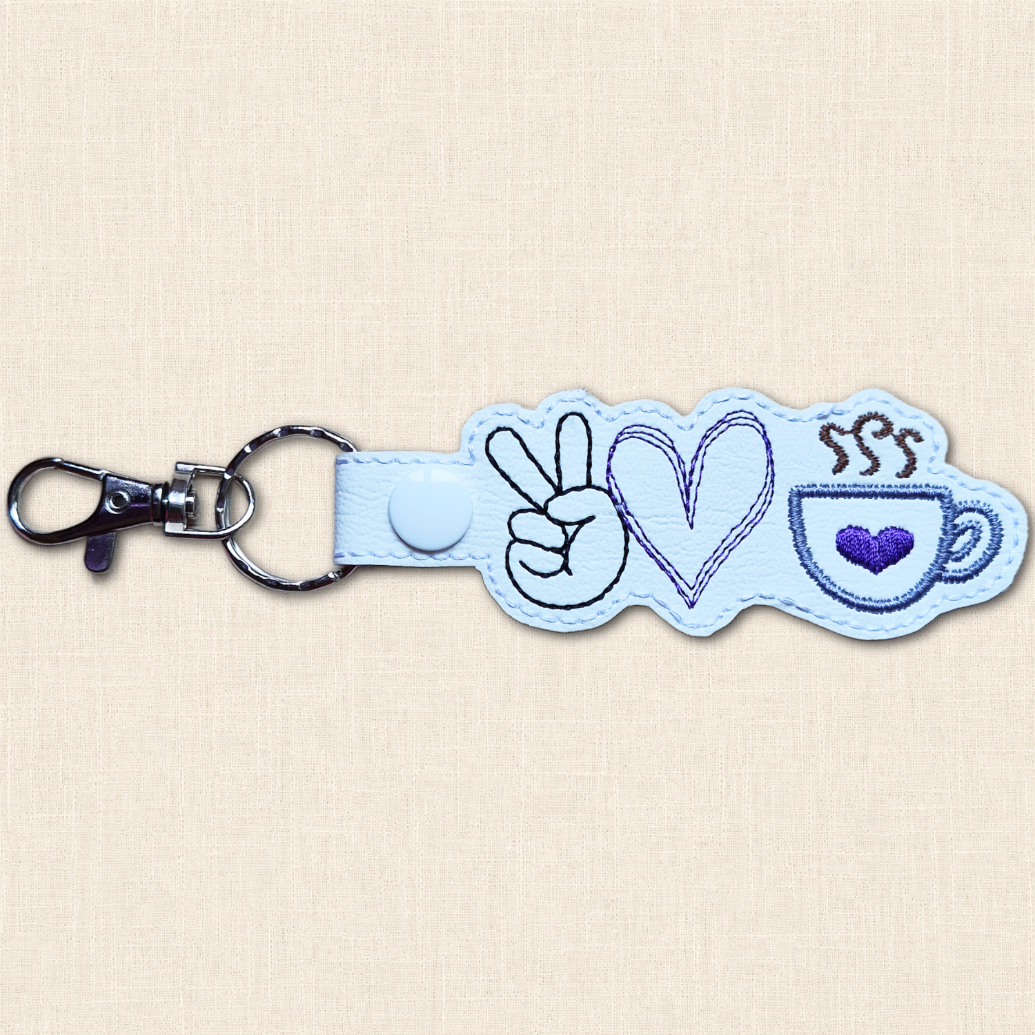 White faux leather keyring with a black peace sign hand, purple outlined heart and blue coffee cup featuring a purple heart and brown steam.