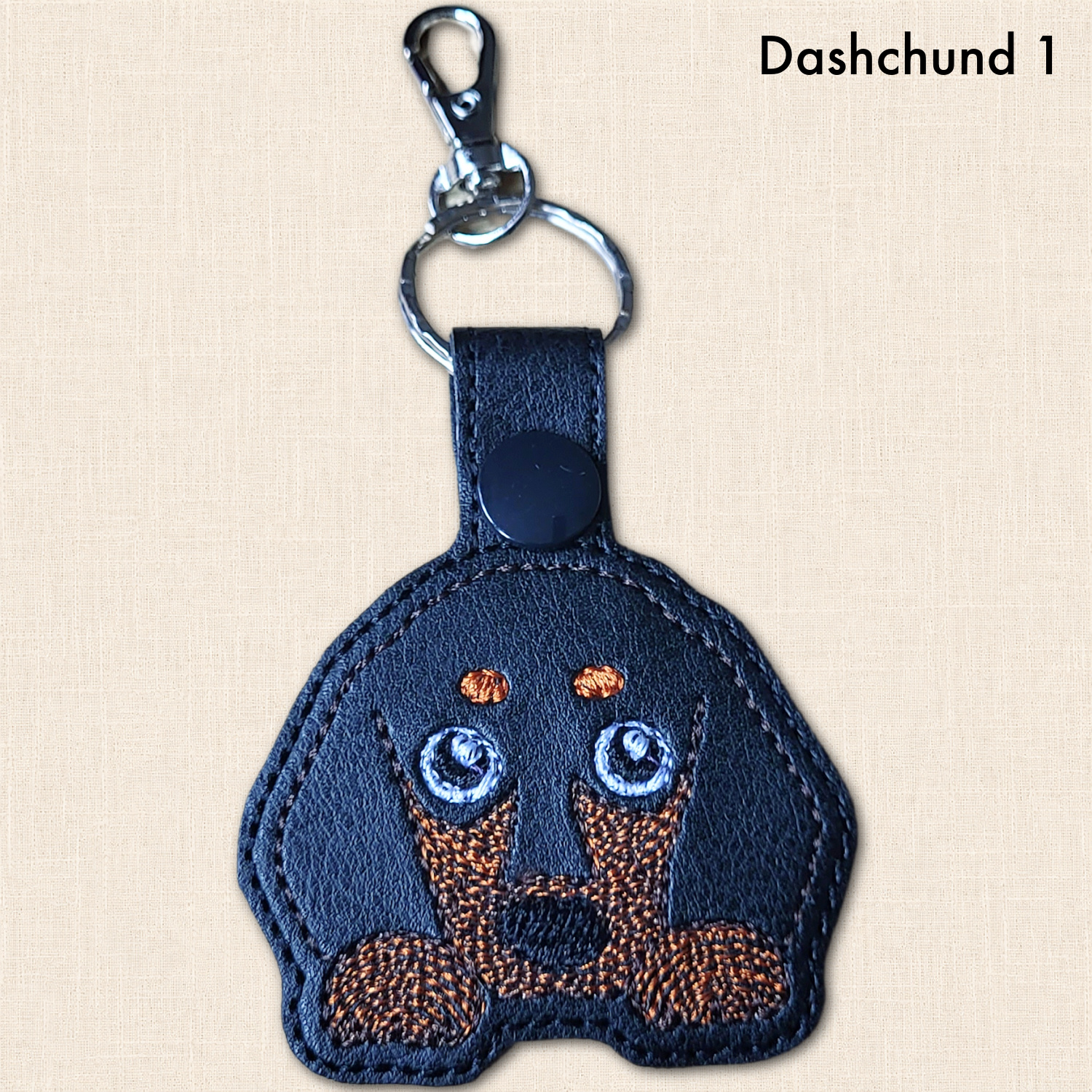 Short haired Dachshund keyring with large embroidered eyes, brown muzzle and peeking paws, labelled ‘Dachshund 1’.