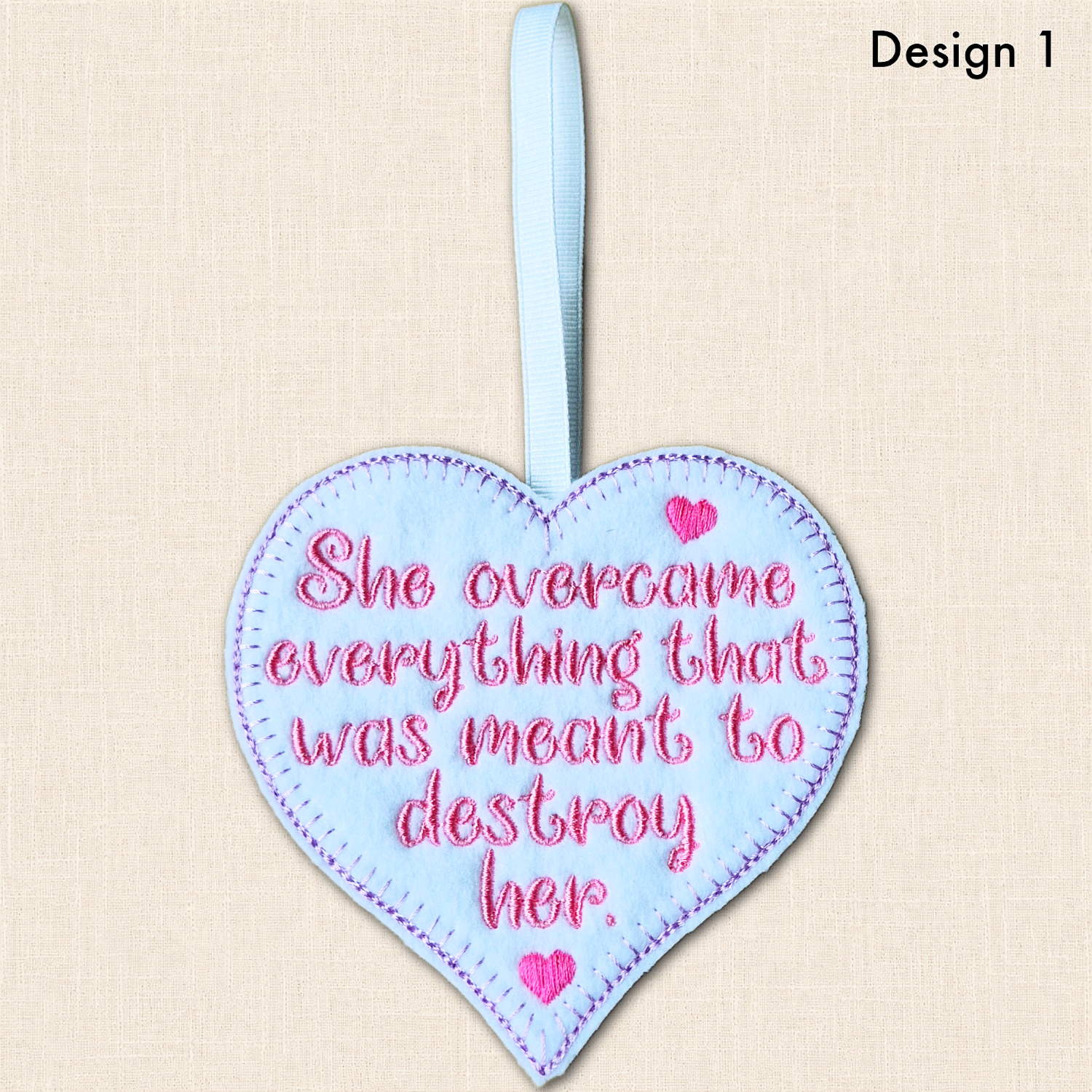Image labelled 'Design 1', showing the resilience heart with pink embroidered wording and hearts, a purple blanket stitch border and white ribbon loop.