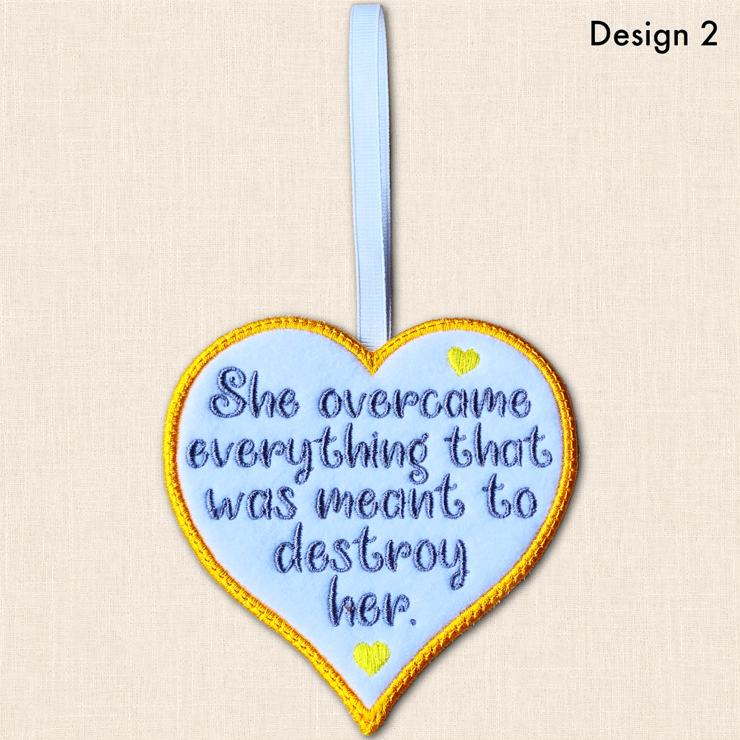 Image labelled 'Design 2', showing the resilience heart with grey wording, yellow hearts, a golden yellow satin stitch border and white ribbon loop.