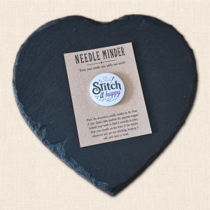 Round white 'Stitch It Happy' needle minder with purple 'Happy' wording and heart detail, presented on a kraft card on a slate heart.