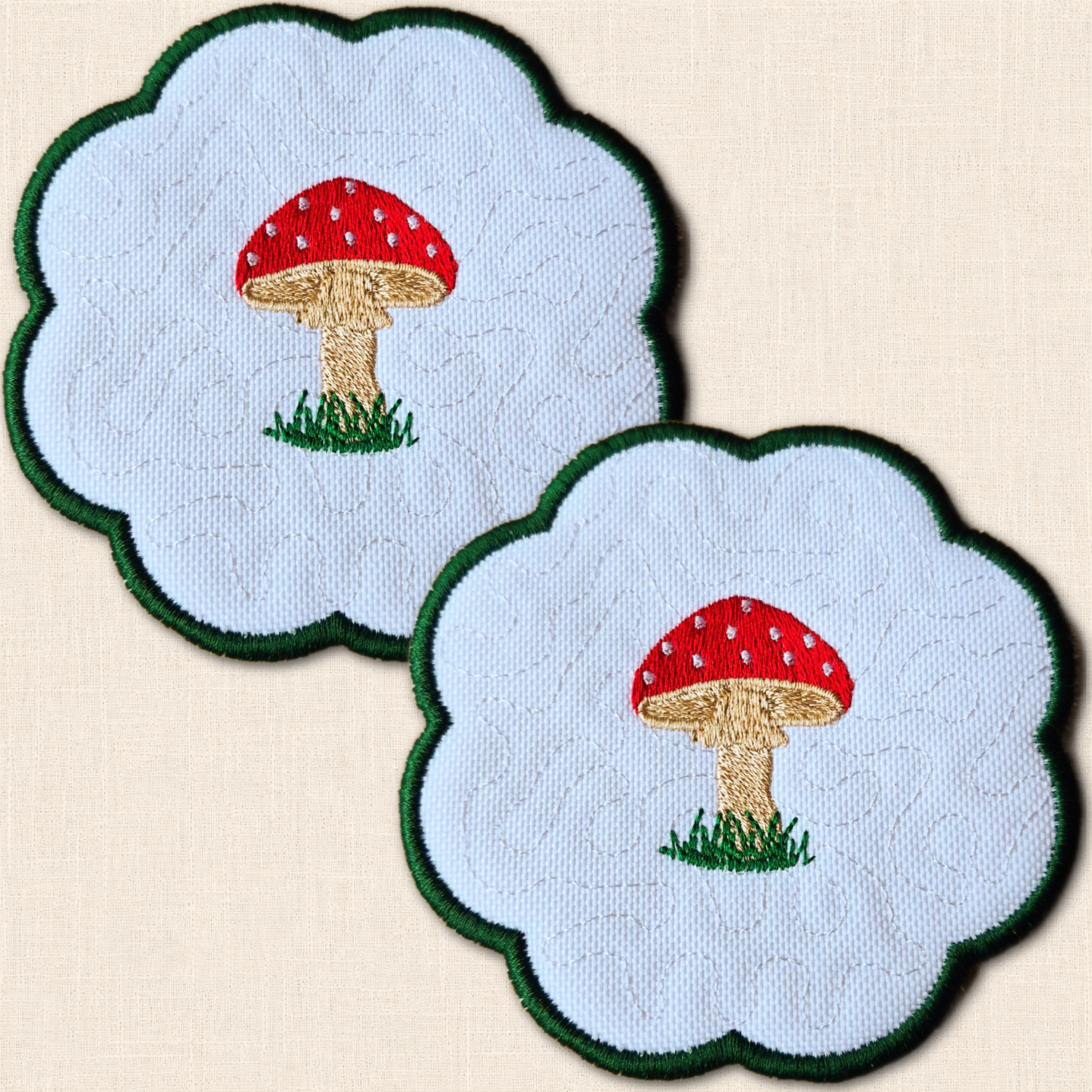 Two white candle coasters with embroidered red and white toadstools, green grass and scalloped green borders.