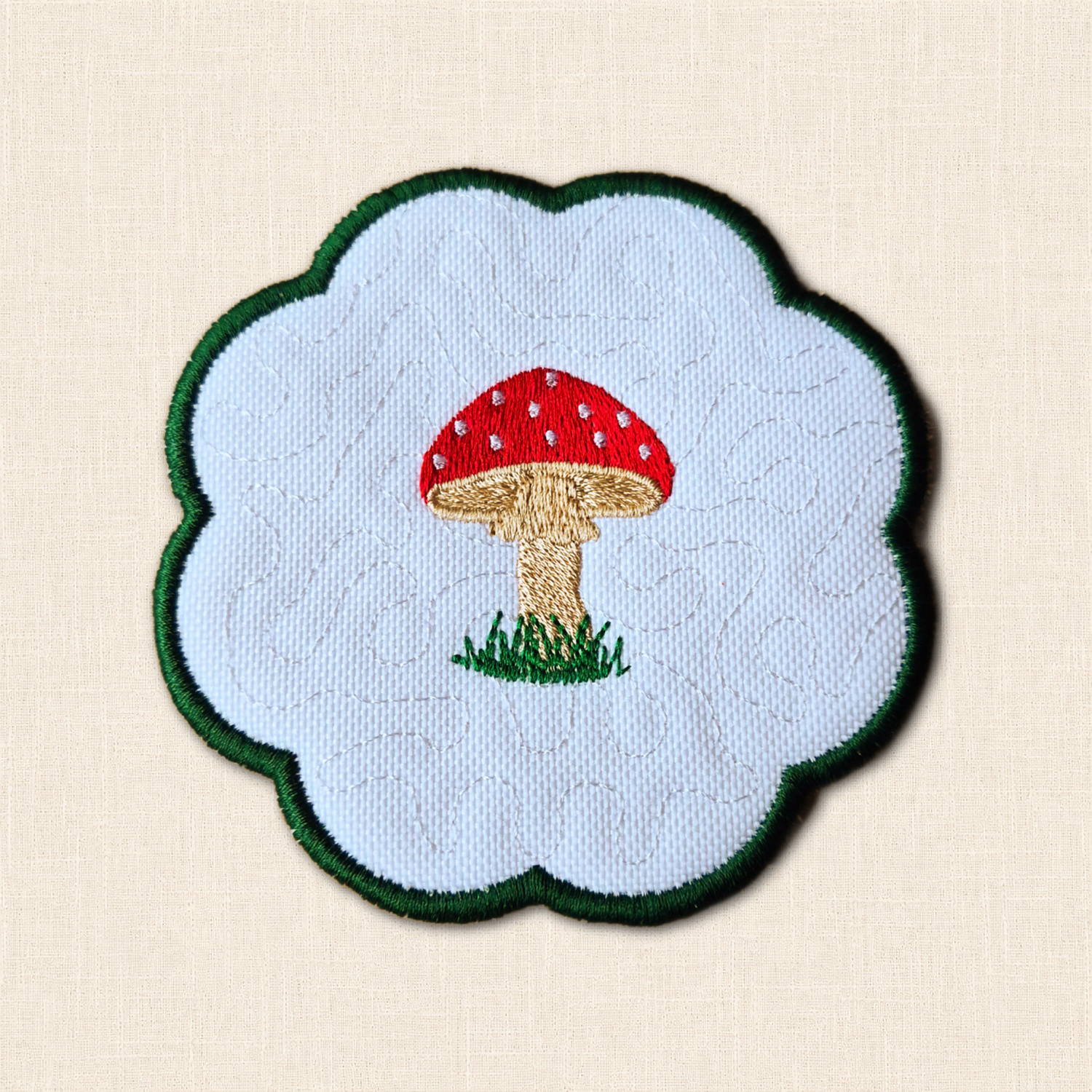 White fabric candle coaster featuring an embroidered red and white toadstool, beige stem, green grass and scalloped green edge.