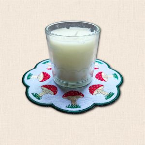 Glass candle jar displayed on a white embroidered toadstool candle mat with red spotted mushrooms and a green scalloped border.