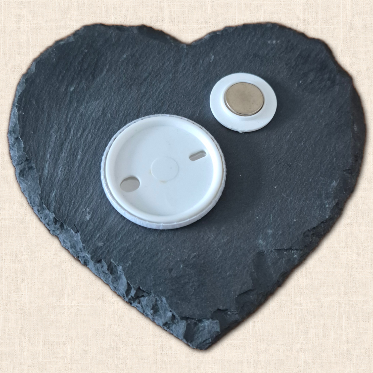 White plastic back of a round needle minder beside its separate backing magnet, displayed on a slate heart.